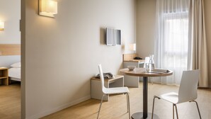 Moderni hotellihuone, jossa on sänky, seinälle asennettu televisio, ruokailutila pöydällä ja tuoleilla sekä suuri ikkuna, jossa on verhot.