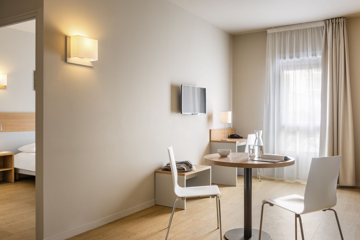 Una habitación de hotel moderna con una cama, un televisor instalado en la pared, una zona de comedor con una mesa y sillas, y un ventanal con cortinas.