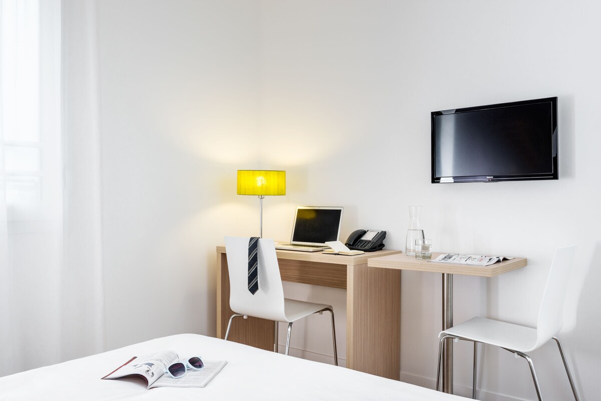 A hotel room with a desk, two chairs, a TV, a lamp, and a bed.