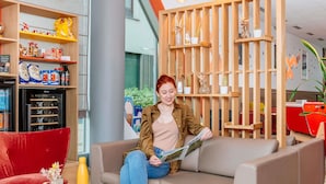 Una mujer sentada en un sofá leyendo una revista en un interior moderno con estanterías de madera y un refrigerador con puerta de vidrio.