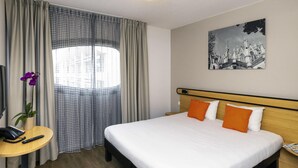 A hotel room with a bed, a desk, a chair, a lamp, a window with curtains, and a wall-mounted picture. - Aparthotel Adagio Access Nancy Centre (Nancy)