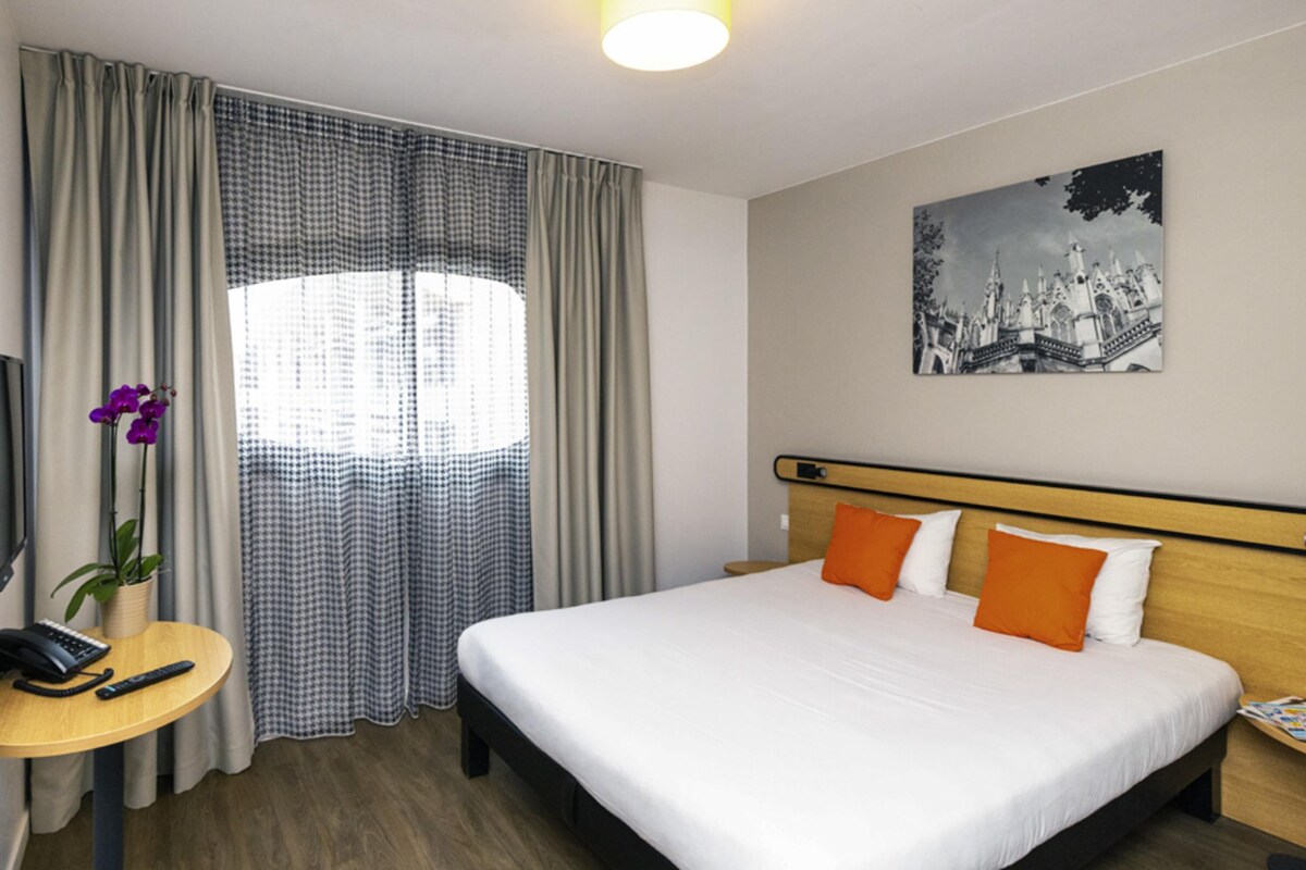 A hotel room with a bed, a desk, a chair, a lamp, a window with curtains, and a wall-mounted picture.