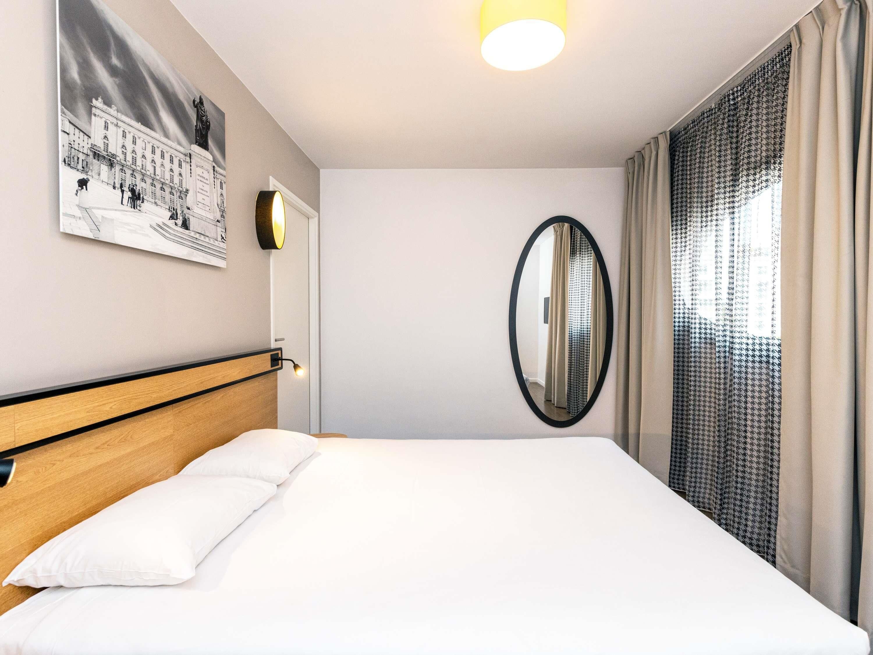 A hotel room with a bed, a wall-mounted mirror, a framed picture, and a window with curtains.