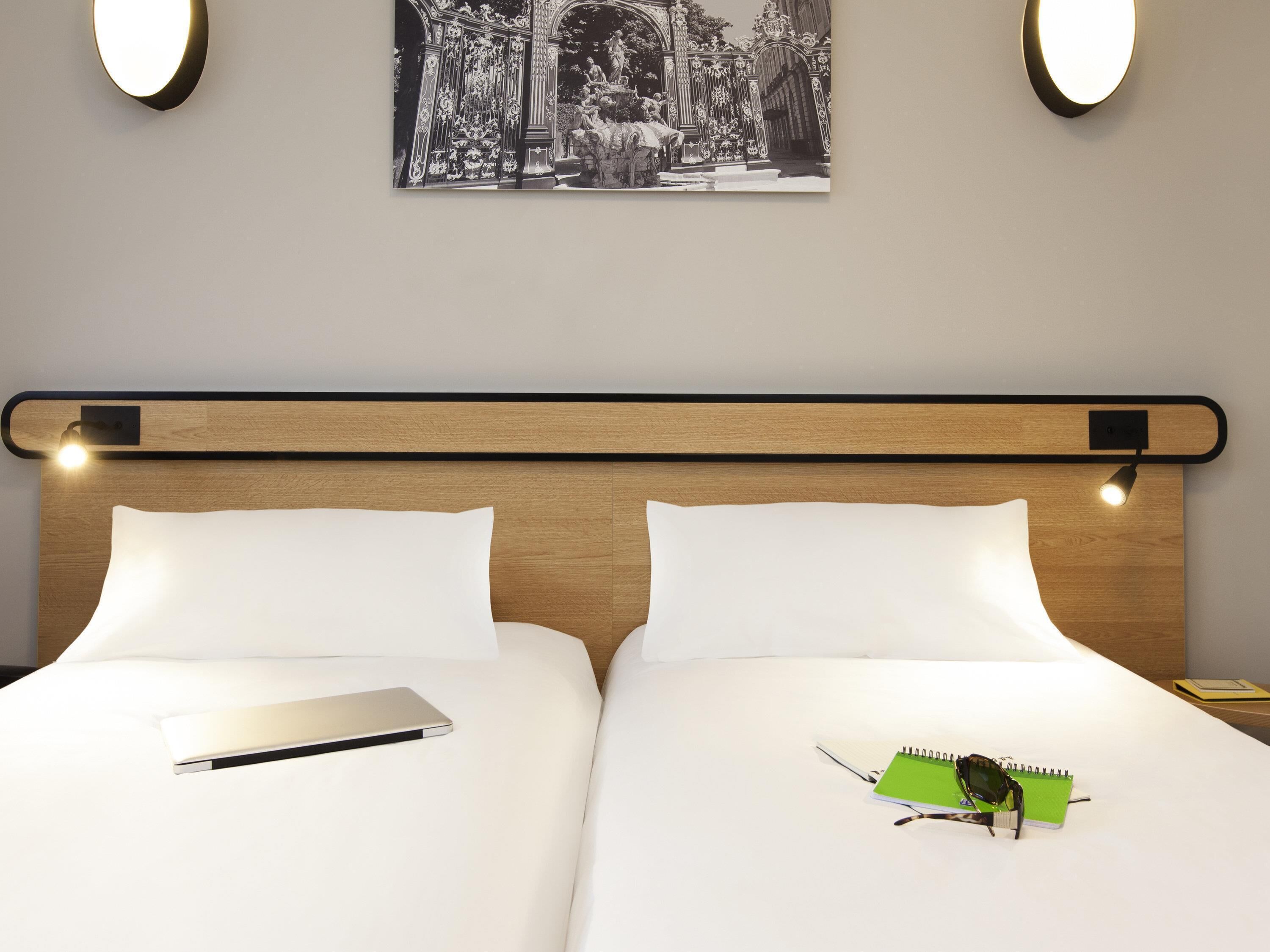 A double bed with a wooden headboard, two bedside lamps, a black and white photo on the wall, and a green notebook with a pen on the bed.