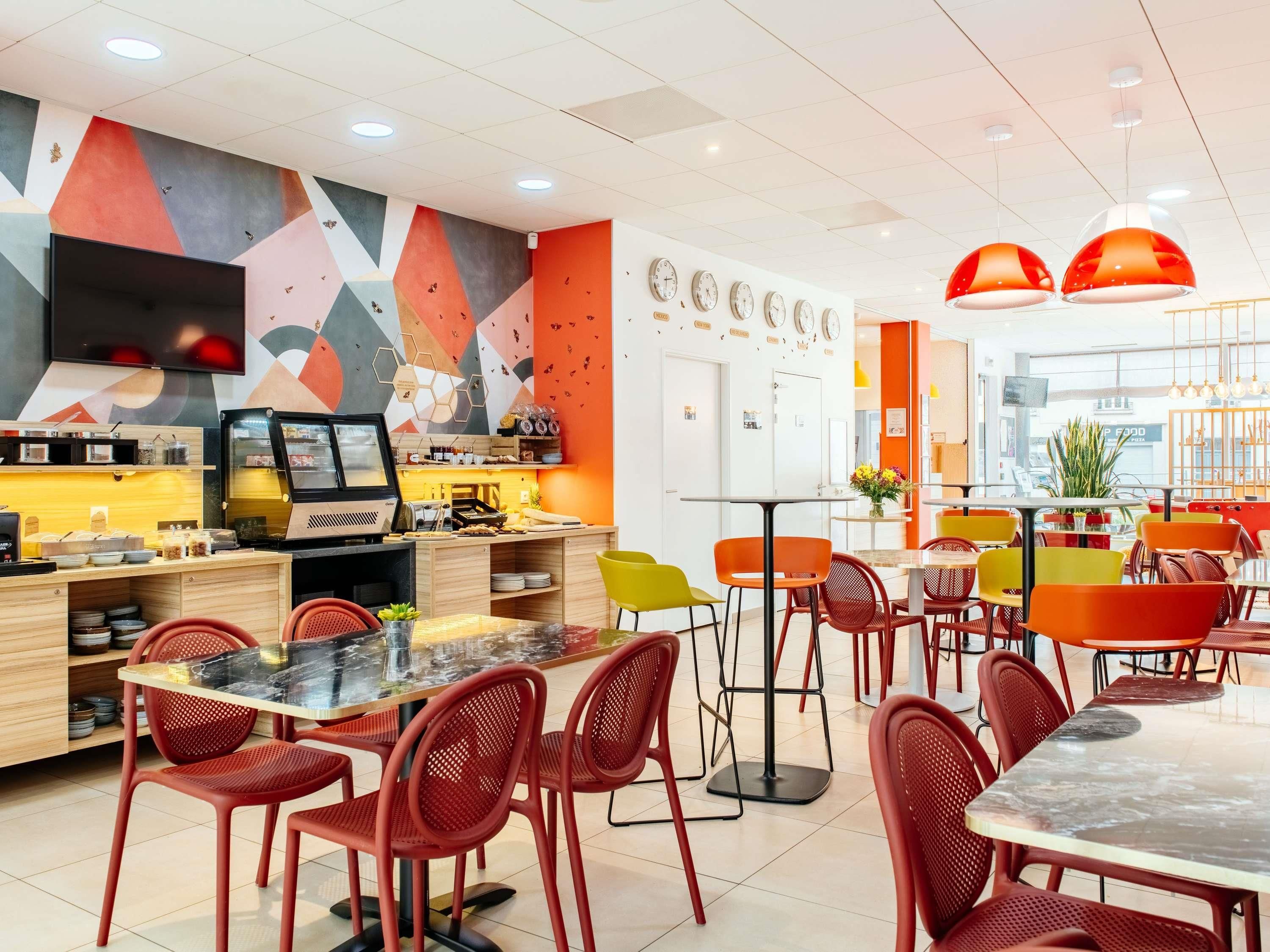 Un café moderne avec une fresque murale géométrique aux couleurs vives, un comptoir en libre-service et des chaises rouges et jaunes.