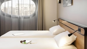 Una habitación de hotel con cama, escritorio, lámpara, ventana con cortinas e imagen colgada en la pared.