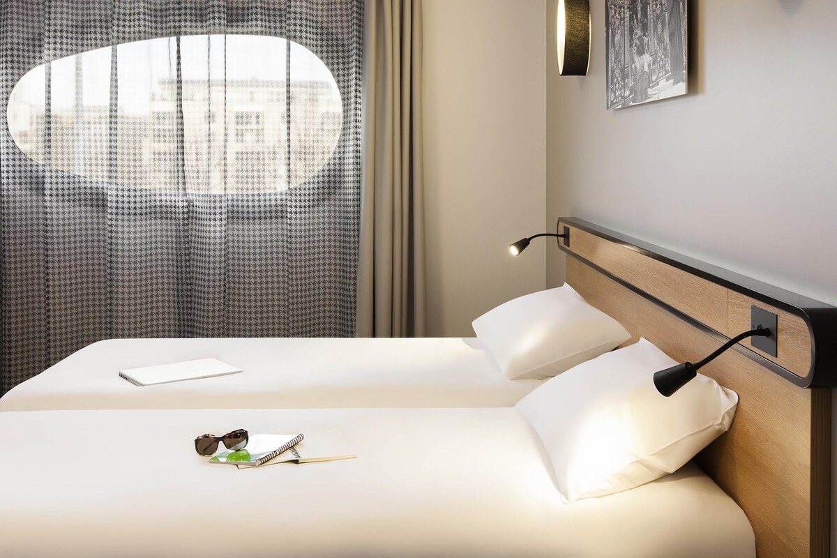 A hotel room with a bed, a desk, a lamp, a window with curtains, and a wall-mounted picture.