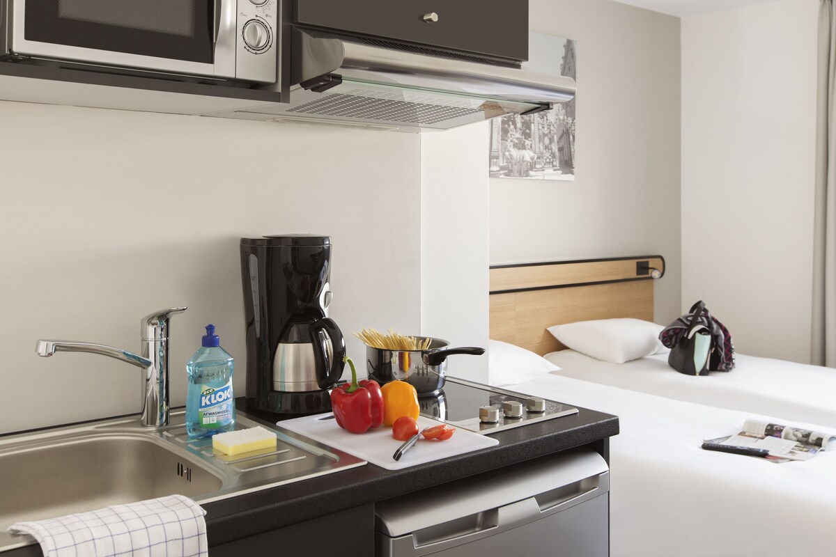 A compact hotel room with a kitchenette, a bed, a bedside table, and a window with a view.