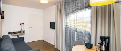 A modern hotel room with a sofa, a small table, a dining table, a coffee maker, and a TV.