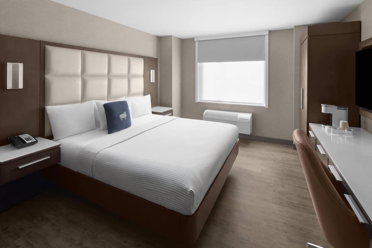 Una habitación de hotel moderna con una cama grande, una mesita de noche con teléfono, un televisor de pantalla plana y una ventana con persianas.