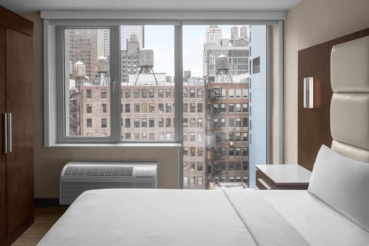 Una habitación de hotel moderna con un ventanal grande, una cama, una mesita de noche y un armario empotrado.
