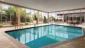 An indoor swimming pool with clear glass walls, a tiled floor, and a seating area with tables and chairs.