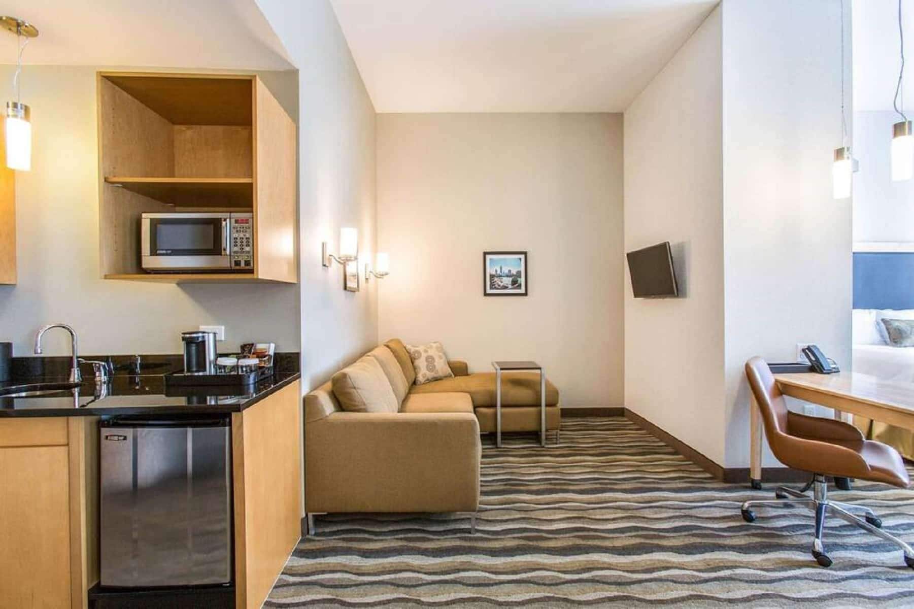 A compact hotel room with a kitchenette, a sofa, a small table, and a desk with a chair.