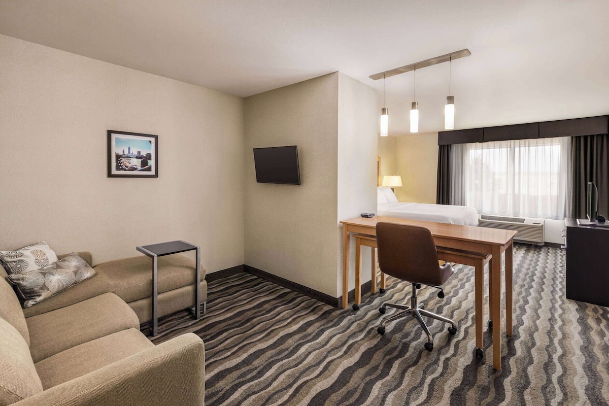 A hotel room with a bed, desk, chair, and a small table. There is a television mounted on the wall and a picture frame.