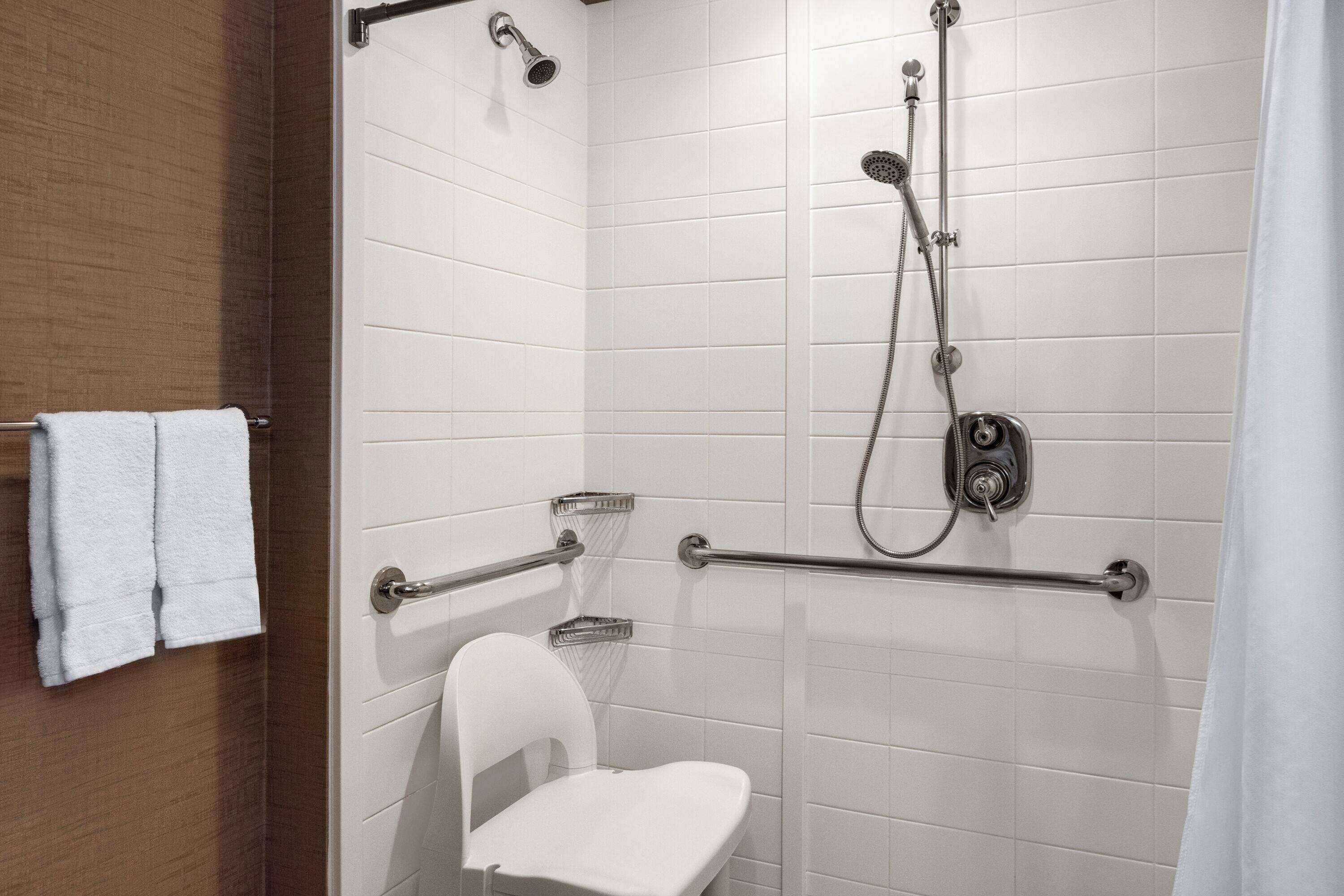 A modern bathroom with white tiled walls, a white toilet, a shower with a handheld showerhead, and grab bars.