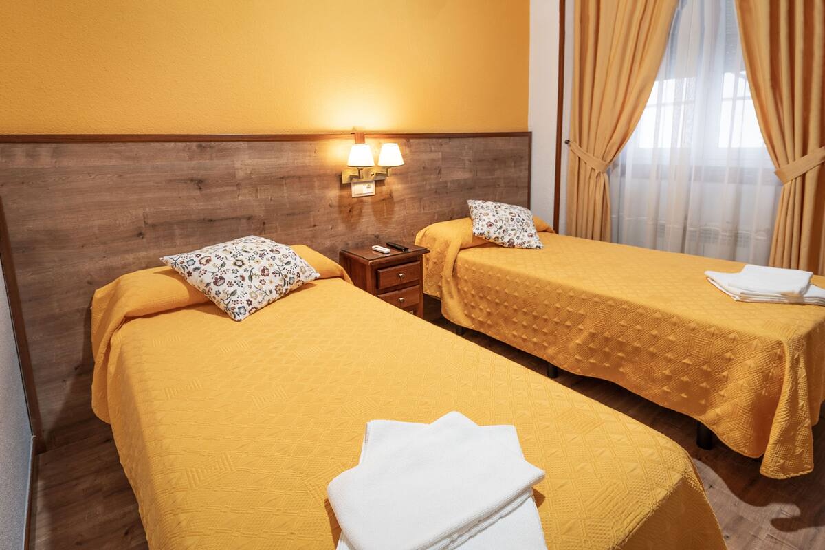 A hotel room with two beds, each with a yellow bedspread and floral pillows. There is a nightstand with a lamp between the beds.