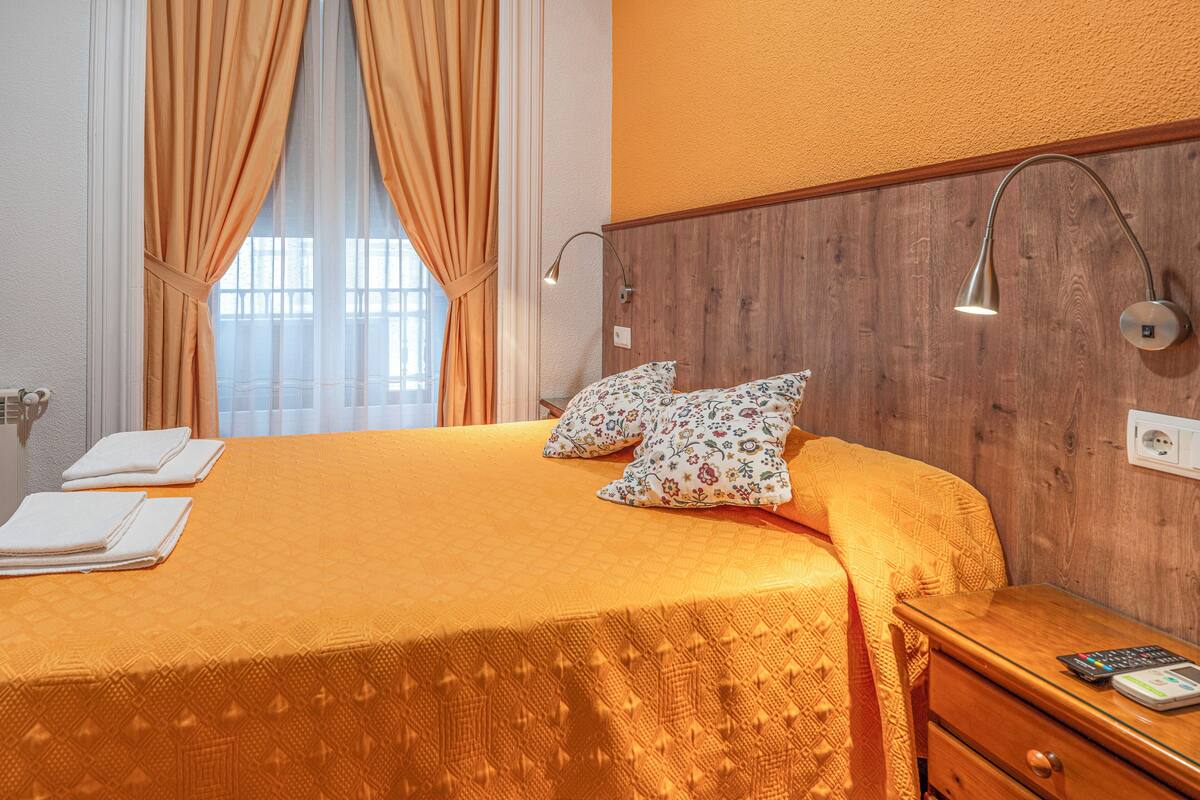A hotel room with a large bed, two pillows, a nightstand, a lamp, and a window with curtains.