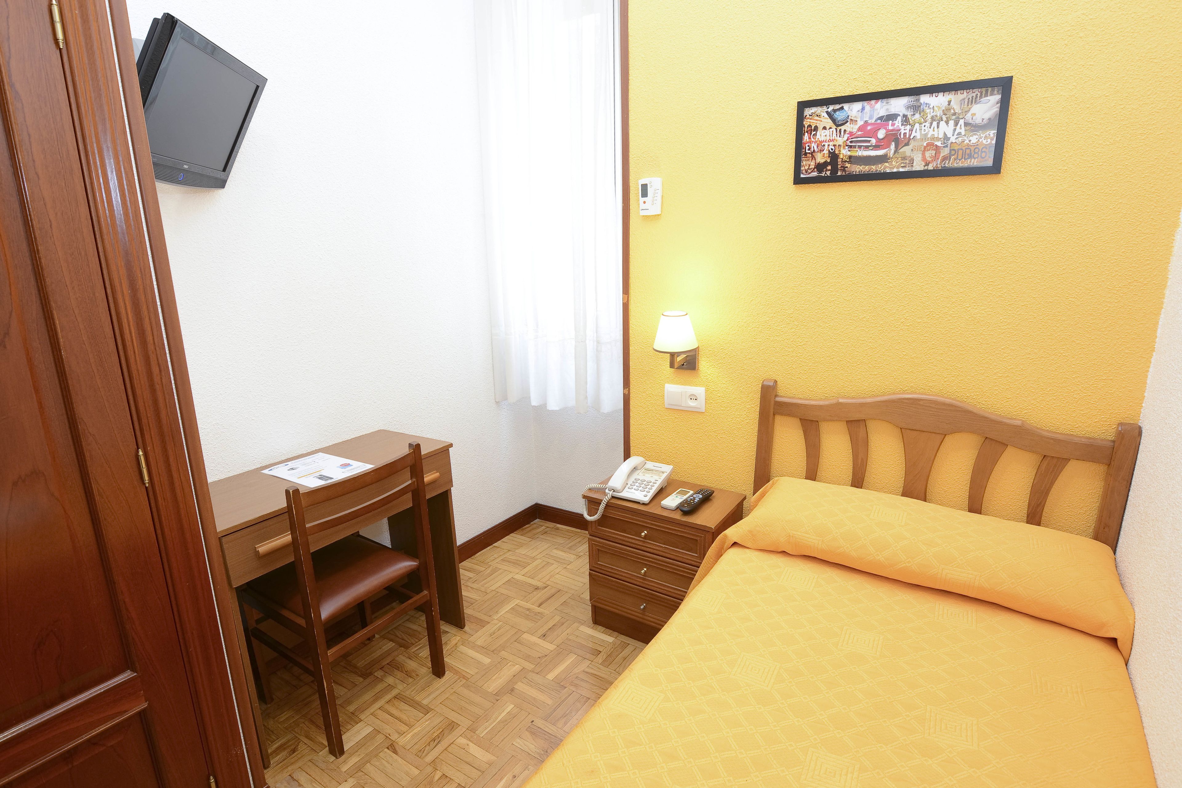 Quarto de hotel com escrivaninha de madeira, cama com colcha laranja, televisão montada e luminária presa à parede.