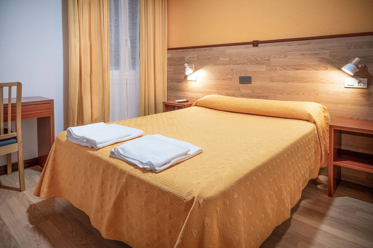 A neatly made bed with a yellow bedspread and two folded white towels, a wooden nightstand with a lamp, and a wooden chair in a hotel room.