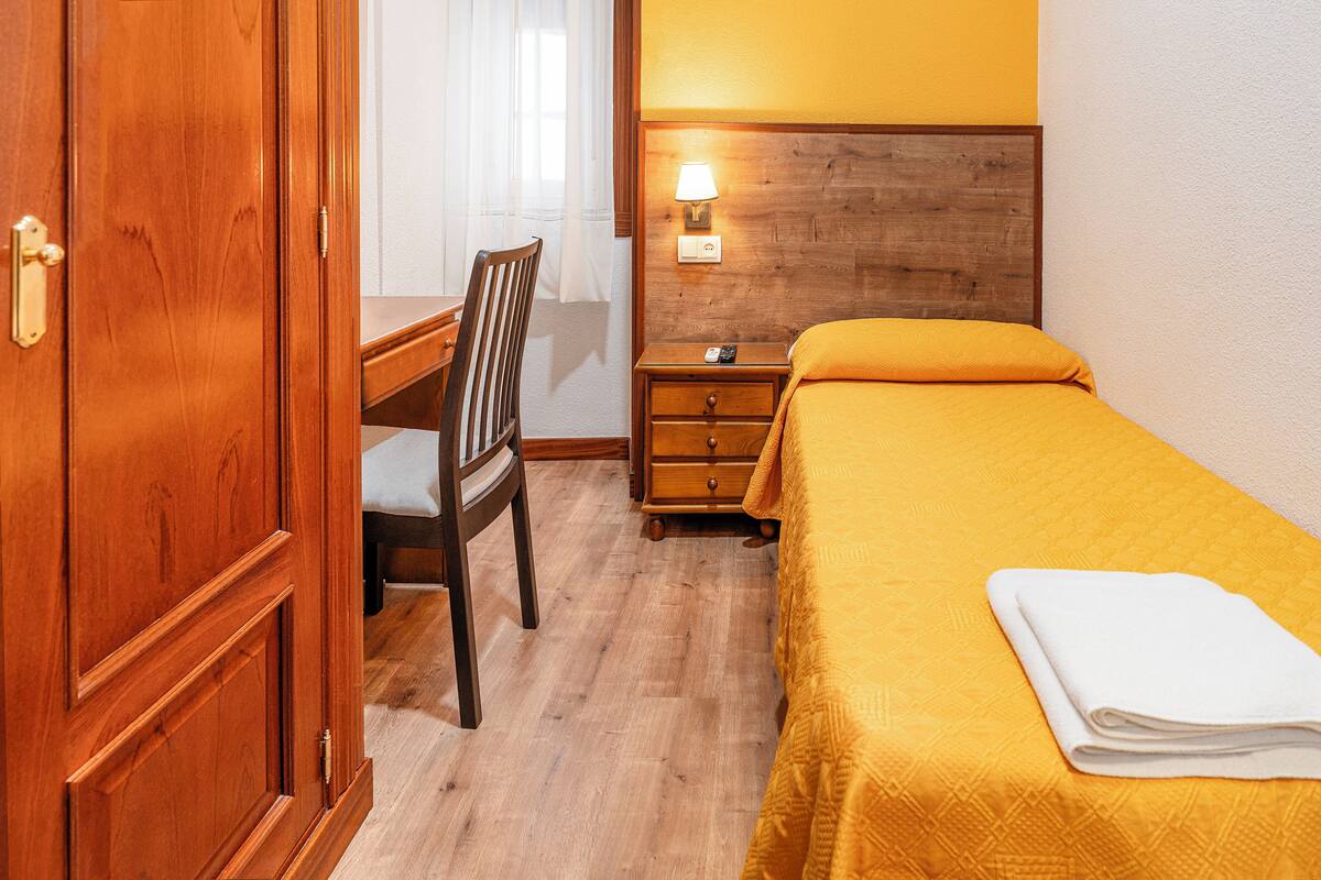 A hotel room with a wooden wardrobe, a desk with a chair, a bedside table with a lamp, and a bed with a yellow bedspread and a folded white towel.