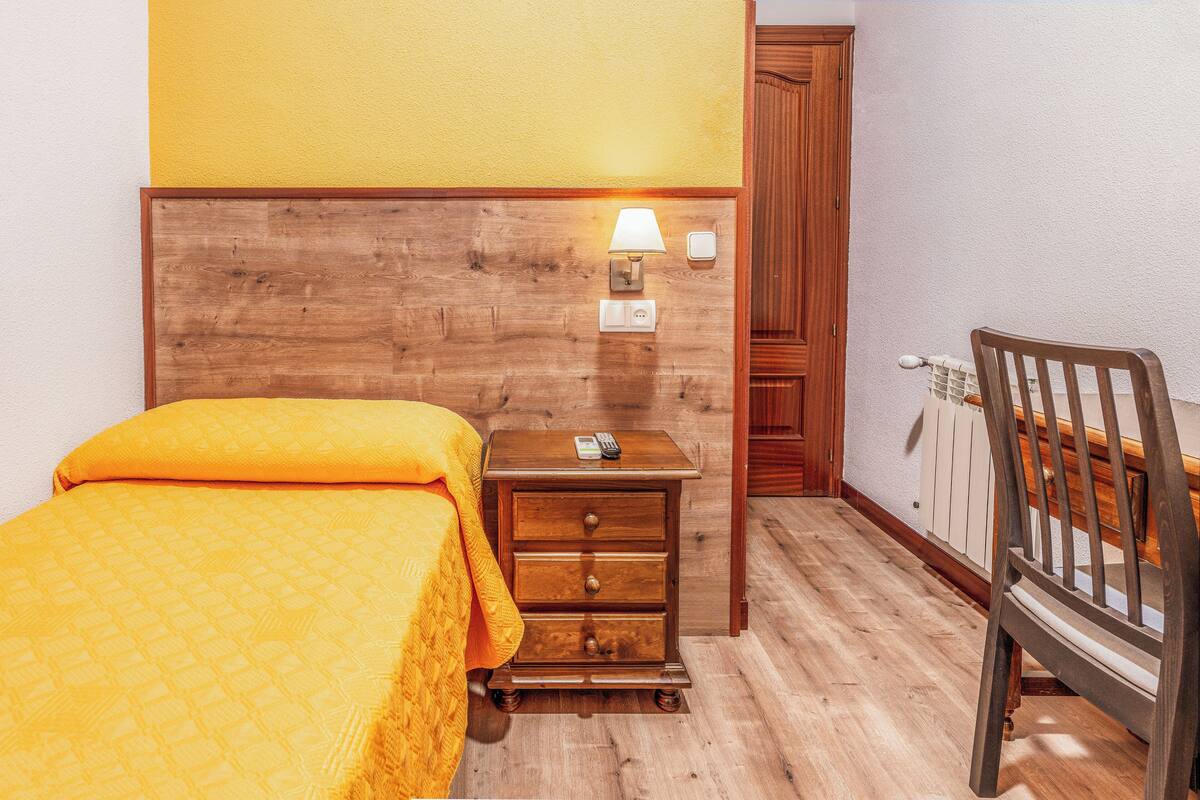A hotel room with a wooden headboard, a bed with an orange bedspread, a nightstand with a lamp, and a wooden chair.