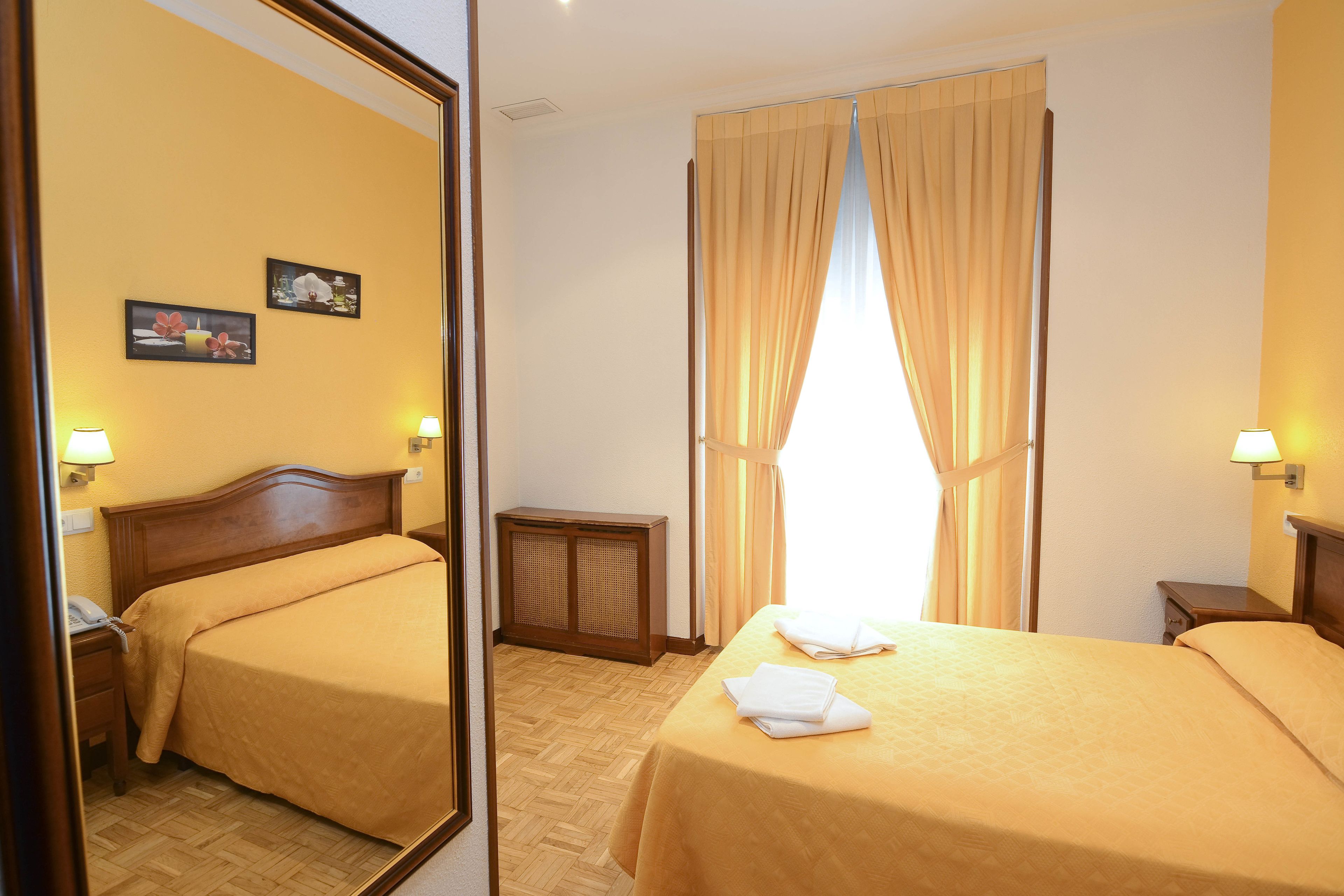 Habitación de hotel con dos camas, cabecero de madera, mesita de noche con lámpara, pequeña mesita de noche con lámpara, teléfono en la pared y ventana con cortinas.