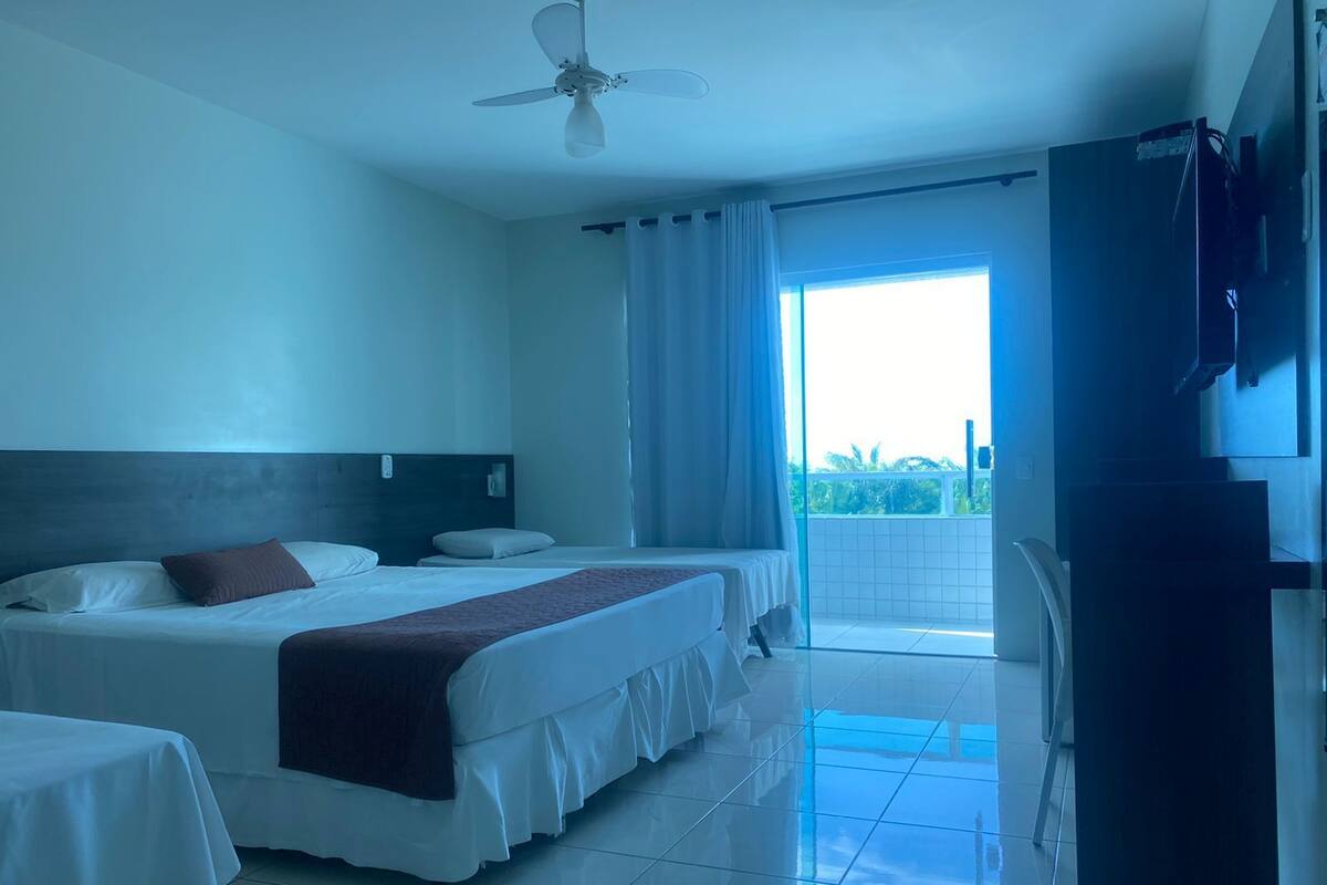 A hotel room with two beds, a ceiling fan, a balcony with curtains, and a TV on the wall.