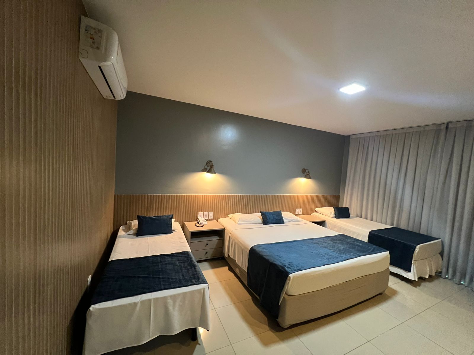 A hotel room with two single beds, a double bed, a bedside table with a lamp, and a large window with curtains.