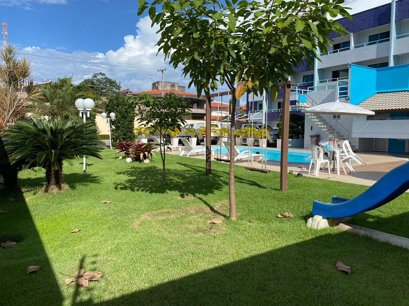 Un área de piscina con sillones de descanso y un tobogán, rodeada de follaje y un edificio con fachada azul y blanca.