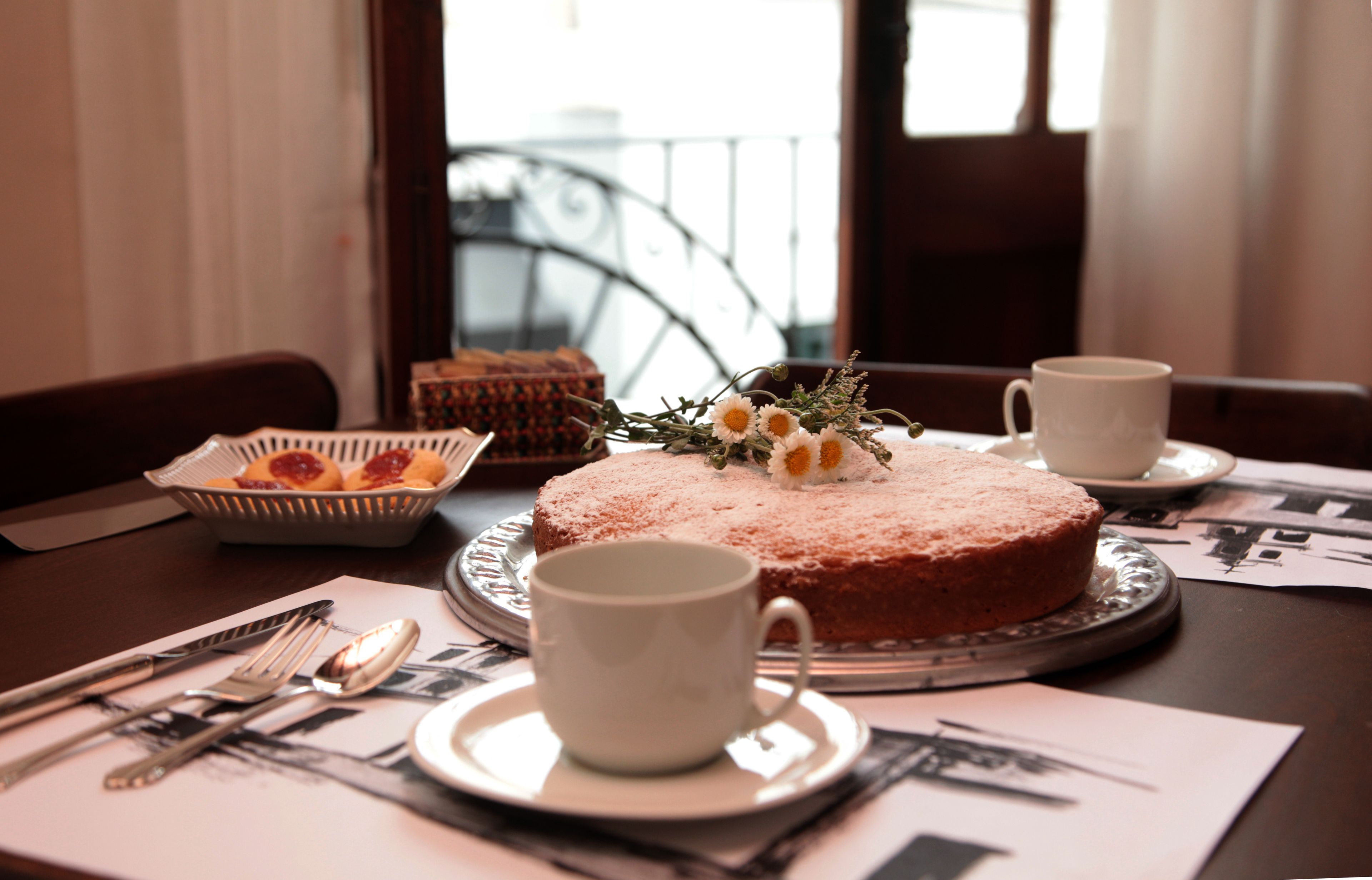A round cake with powdered sugar and a floral decoration on a silver cake stand, accompanied by two white cups and a basket of pastries on a table.