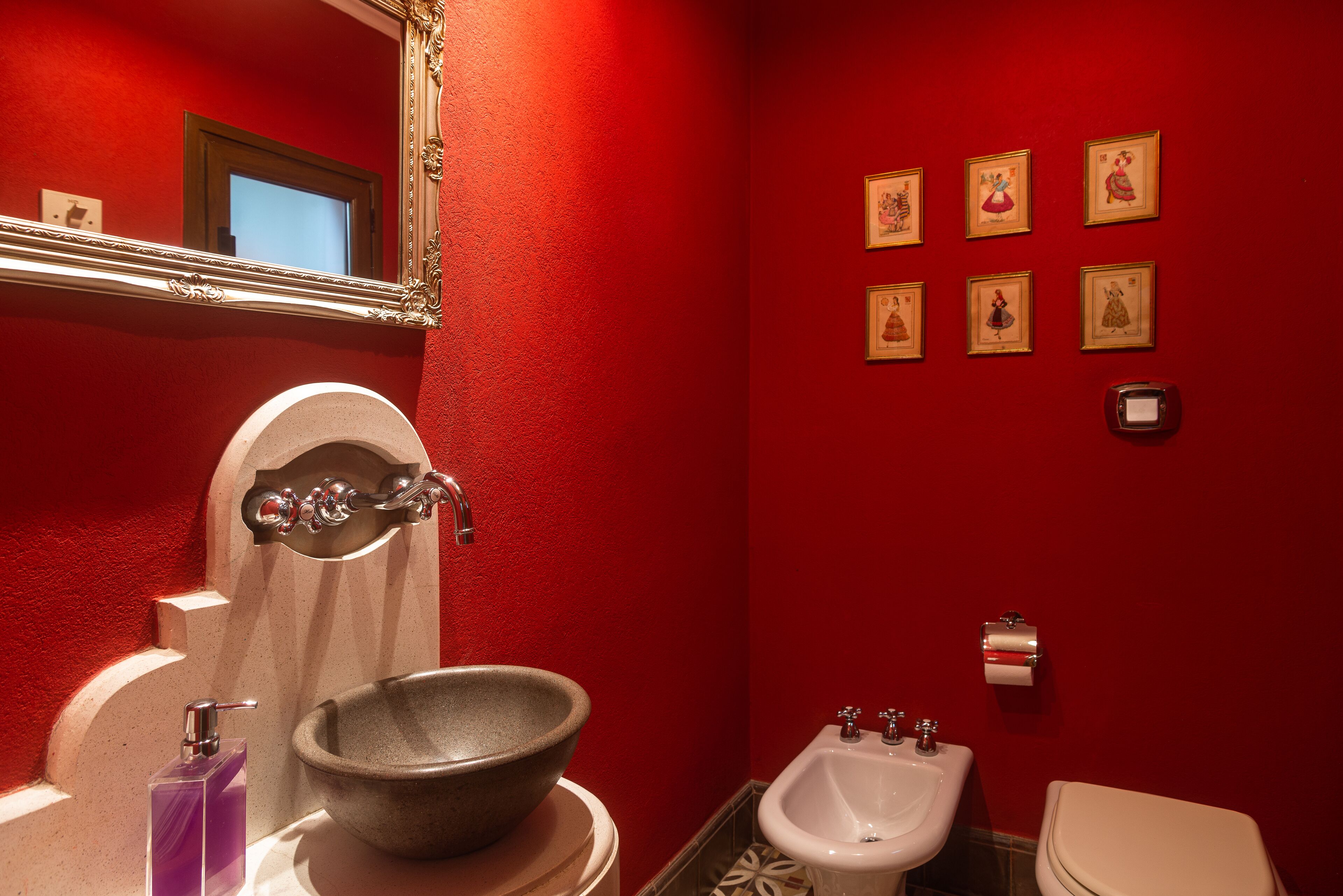 A bathroom with a red wall, a large mirror, a stone sink, a bidet, and several framed pictures.