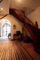 Un pasillo amplio con una escalera de madera, una puerta con detalles de hierro forjado y una alfombra en el suelo.