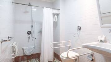 A bathroom with white tiled walls, a shower with a glass door, a toilet with a safety rail, and a sink with a mirror above it.