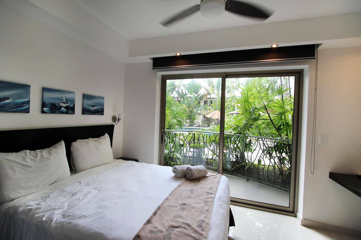 A hotel room with a bed, a ceiling fan, and a large window with a view of greenery.