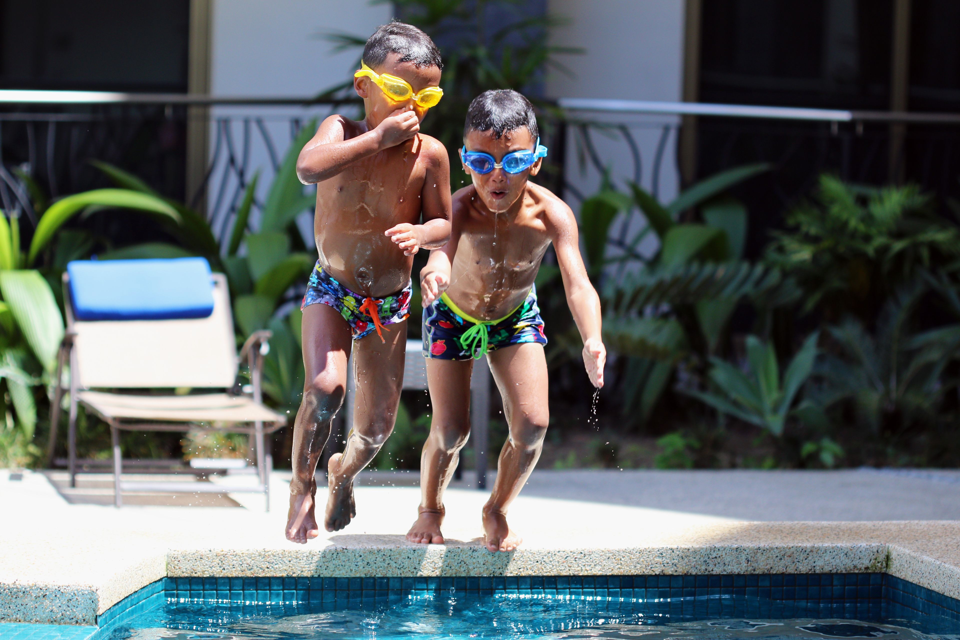 Dos niños saltando a una piscina, con una silla y plantas al fondo.