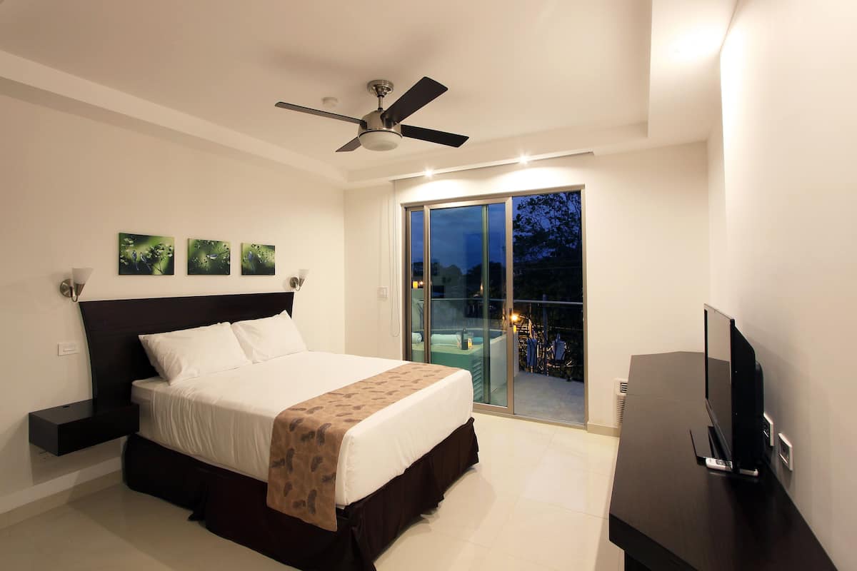A bedroom with a bed, a ceiling fan, a TV, and a balcony with a glass door.