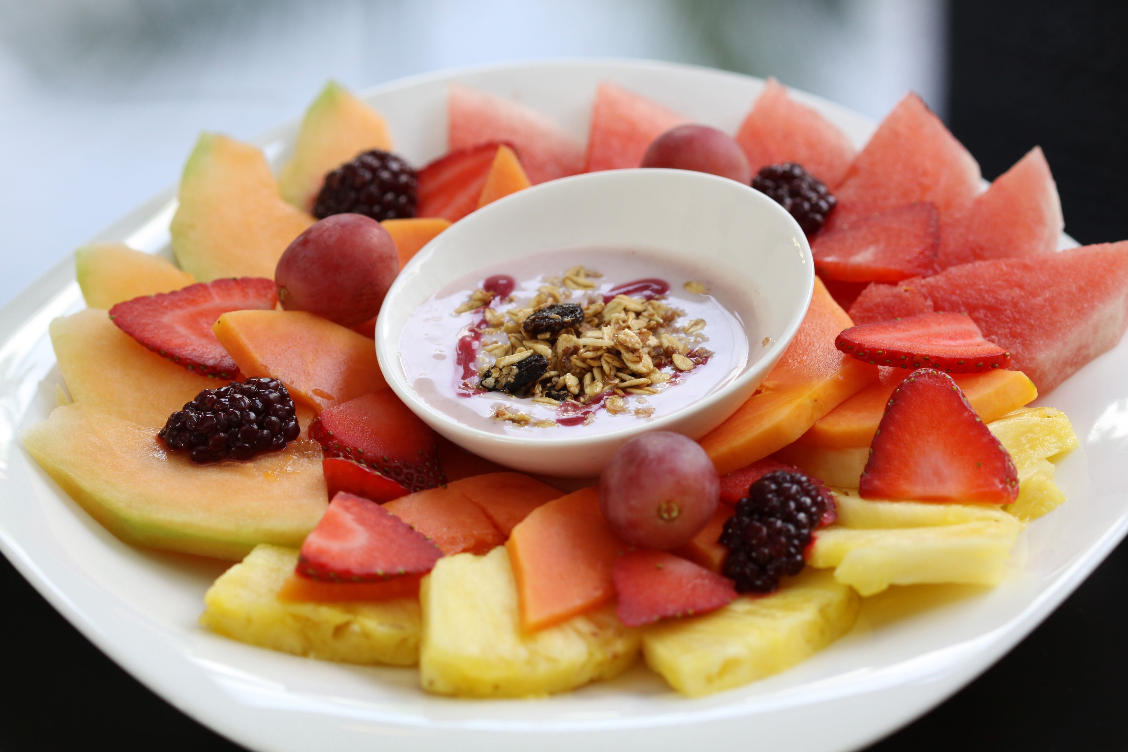 Een fruitschaal met een kom yoghurt en granola in het midden.