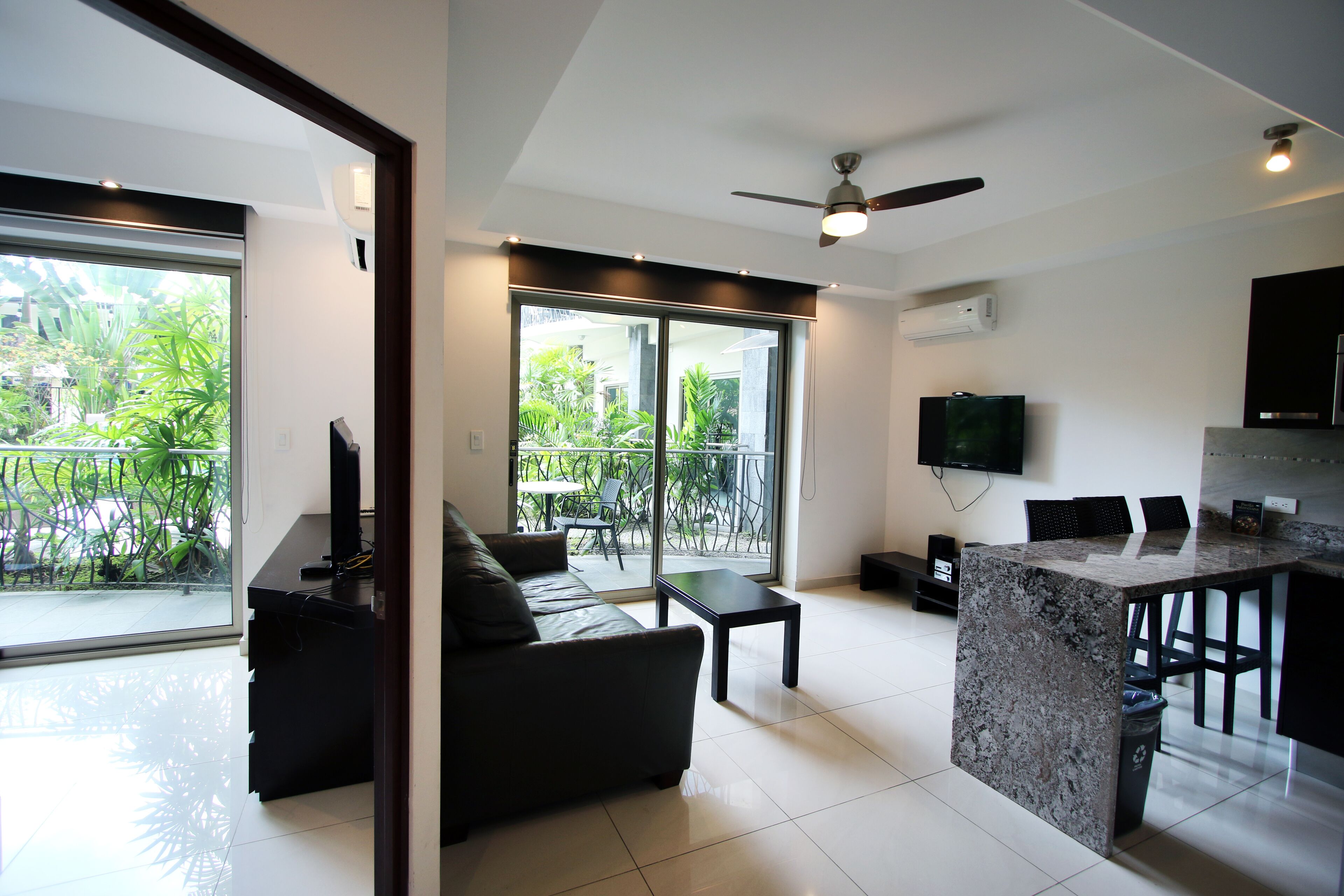 A modern living room with a sofa, a coffee table, and a TV. There is a balcony with plants visible through the glass doors.