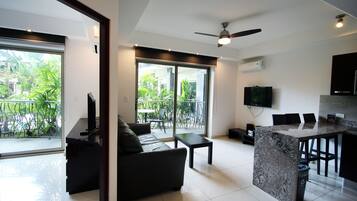 A modern living room with a sofa, a coffee table, and a TV. There is a balcony with plants visible through the glass doors.