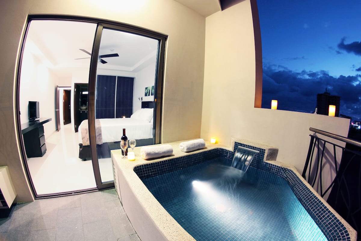 A modern hotel room with a private balcony, a large window, and a view of the city skyline at dusk.