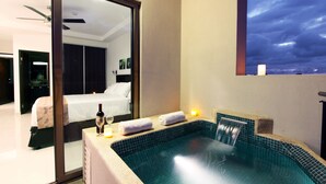 A modern hotel room with a large bed, a small sitting area, a bathtub, and a view of the city at night.