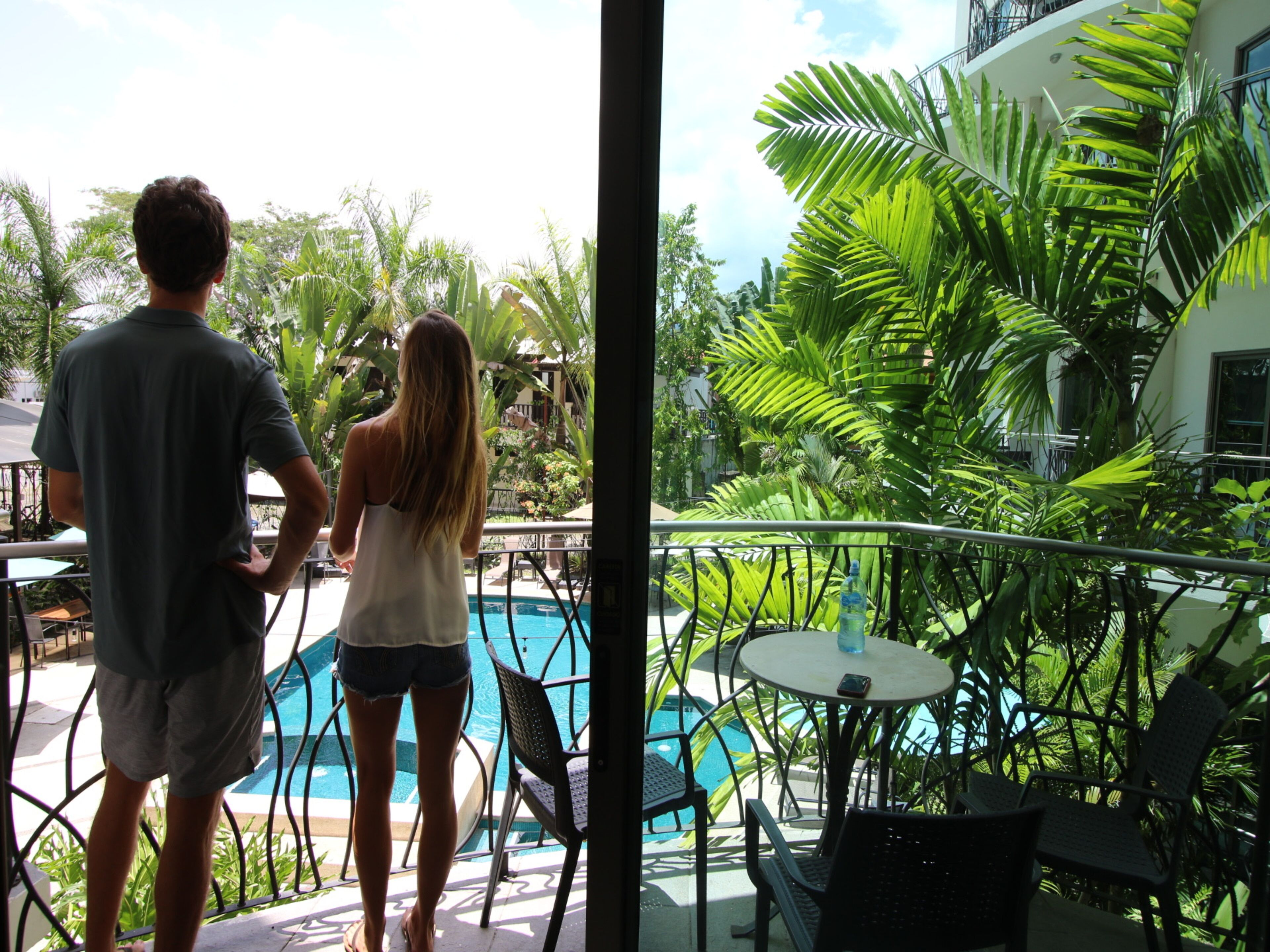 Un hombre y una mujer están junto a una piscina, con vistas a un jardín exuberante y un edificio al fondo.