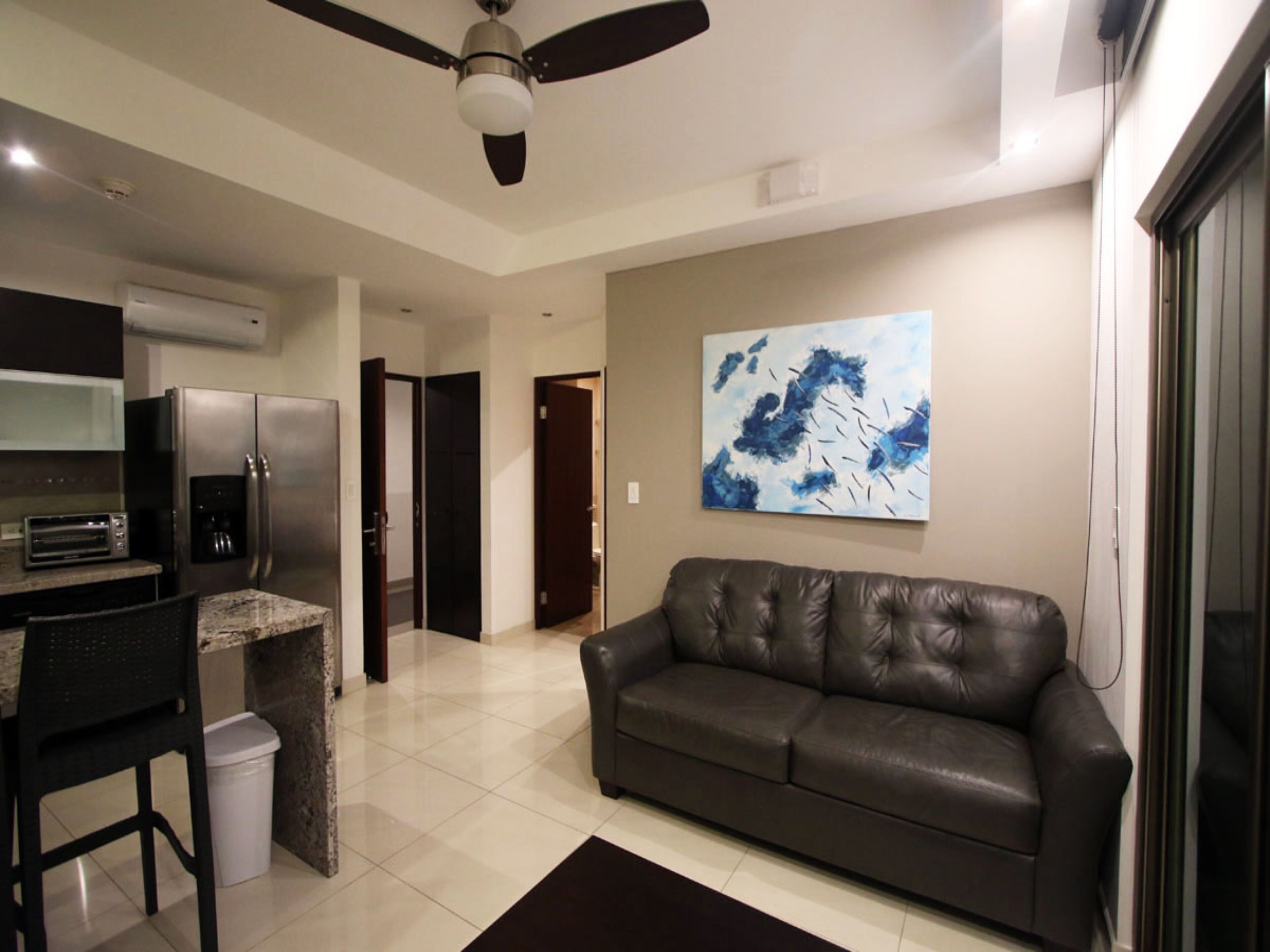 A modern living room with a leather sofa, a dining area with a kitchen in the background, and a ceiling fan.