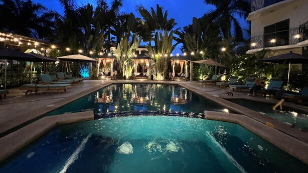 A pool area with lounge chairs and umbrellas, surrounded by palm trees and a building with balconies.