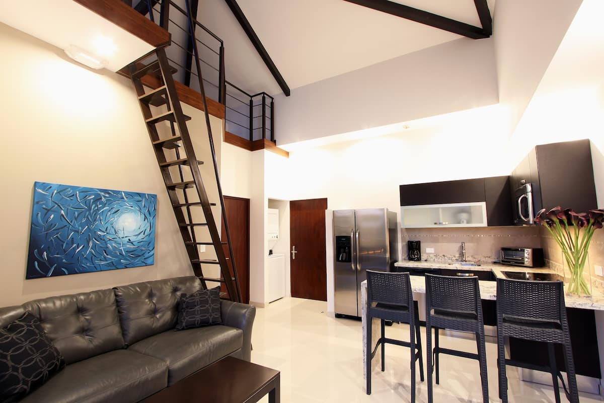 A modern kitchen with a stainless steel refrigerator, microwave, and coffee maker. There is a dining area with a black table and chairs. A staircase leads to an upper level.