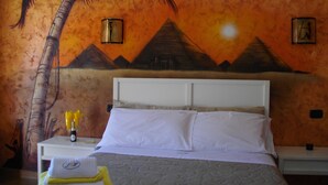 Ett hotellrum med en säng, en vit sänggavel och en väggmålning som föreställer ett palmträd och pyramider.