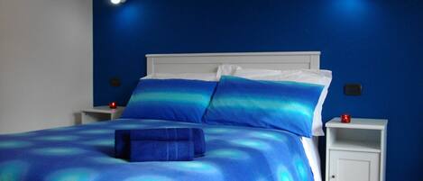 Una cama con ropa de cama azul, cabecera blanca y dos mesitas de noche con velas rojas.