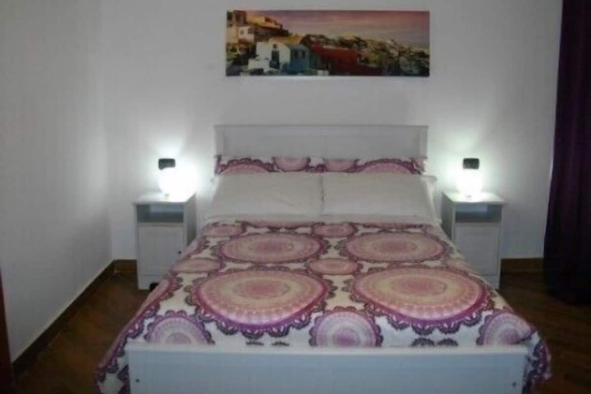Une chambre à coucher comprenant un lit, deux tables de chevet, une commode, un tableau accroché au mur et une fenêtre avec des rideaux.