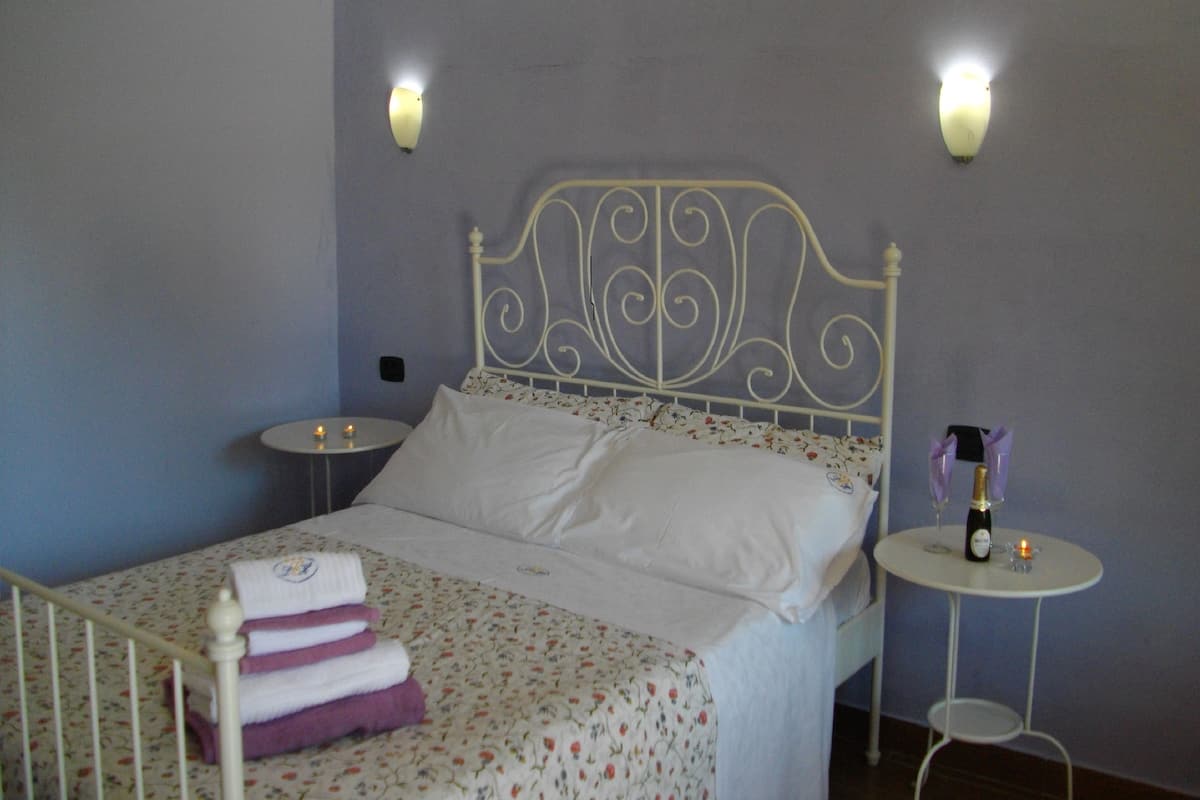 Un lit bien fait, recouvert d’une couvre-lit à motifs floraux, avec des oreillers blancs et des serviettes pliées posées sur une table de chevet. On y trouve également deux autres tables de chevet ornées de bougies et d’une bouteille de champagne.