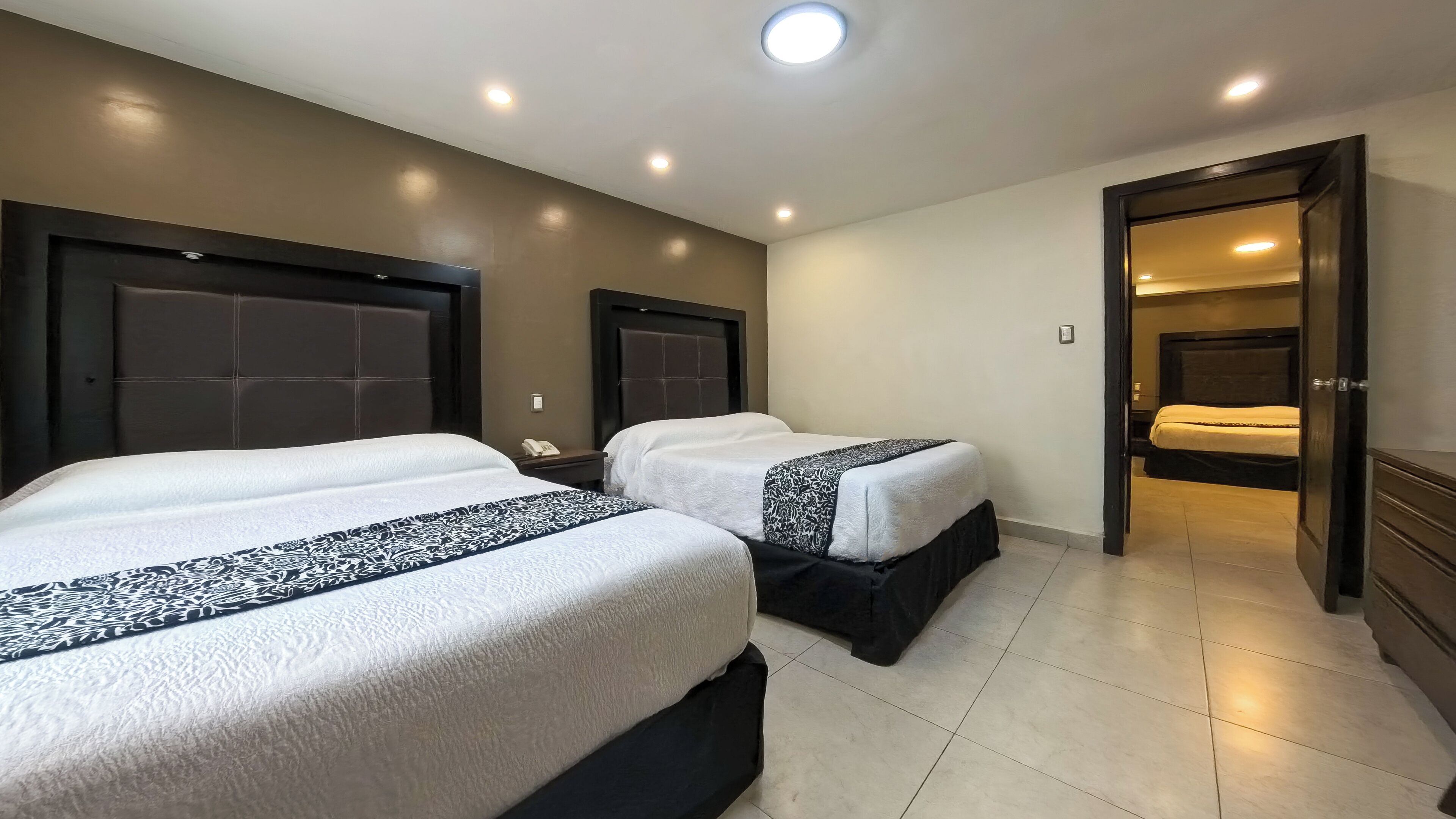 A hotel room with two beds, a dark headboard, a bedside table with a phone, and a door leading to another room.
